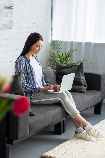 Vista lateral da jovem mulher usando laptop no sofá em casa — Fotografia de Stock