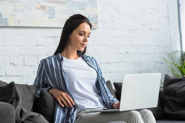 Young focused woman using laptop on sofa at home — Stock Photo