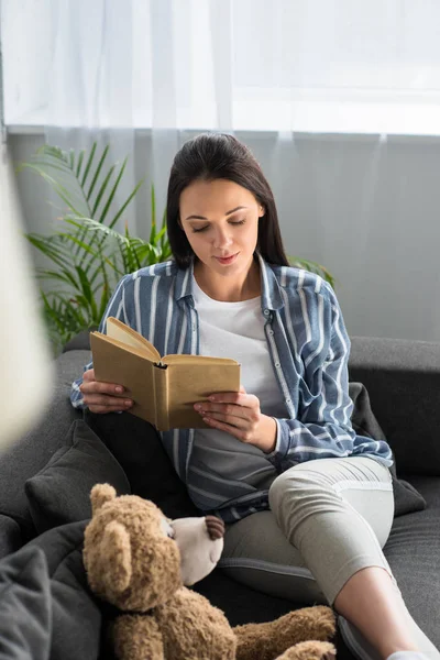 Young woman reading book on sofa with teddy bear at home — Stock Photo