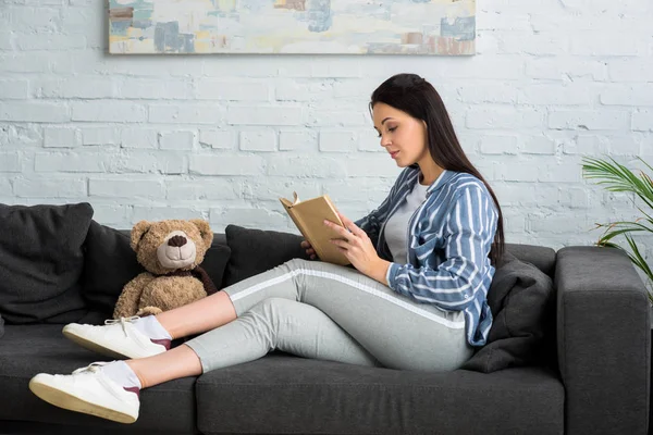 Side view of young woman reading book on sofa with teddy bear at home — Stock Photo