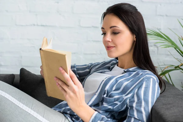 Side view of young woman reading book on sofa at home — Stock Photo