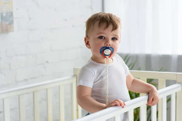 Menino pequeno bonito com chupeta no berço do bebê olhando para a câmera em casa — Fotografia de Stock