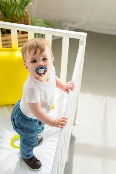 High angle view of cute little baby boy with pacifier in baby crib looking at camera at home — Stock Photo