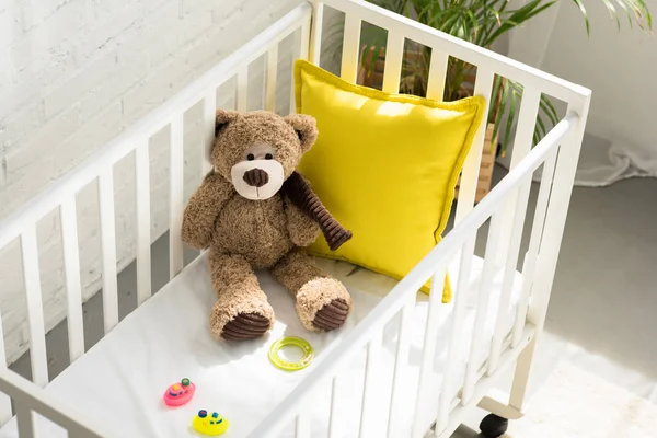 Vista de ángulo alto del oso de peluche, otros juguetes y almohada amarilla en la cuna del bebé en casa - foto de stock