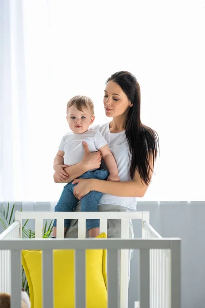 Retrato de la madre con el pequeño bebé en las manos de pie en la cuna del bebé en casa - foto de stock