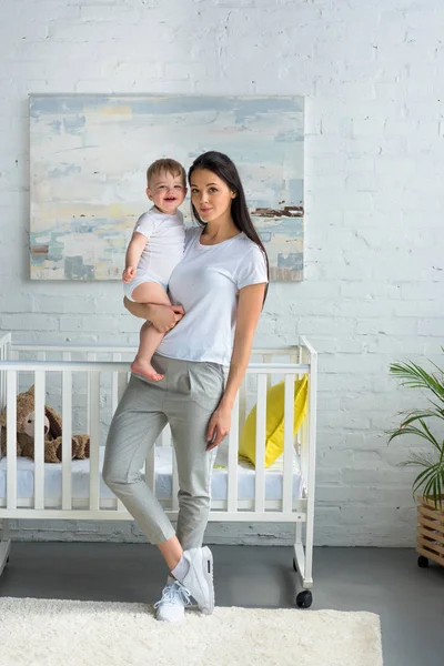 Mother holding cute smiling baby in hands while standing at baby crib at home — Stock Photo