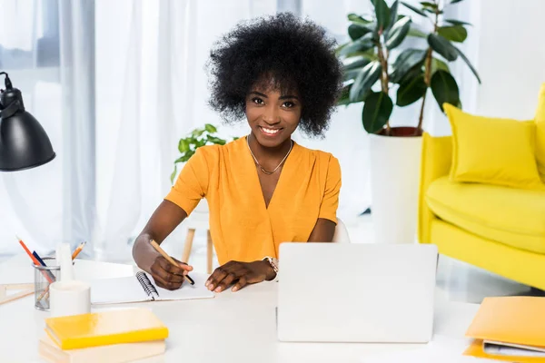 Portrait of smiling african american freelancer at workplace with laptop and notebook at home — Stock Photo