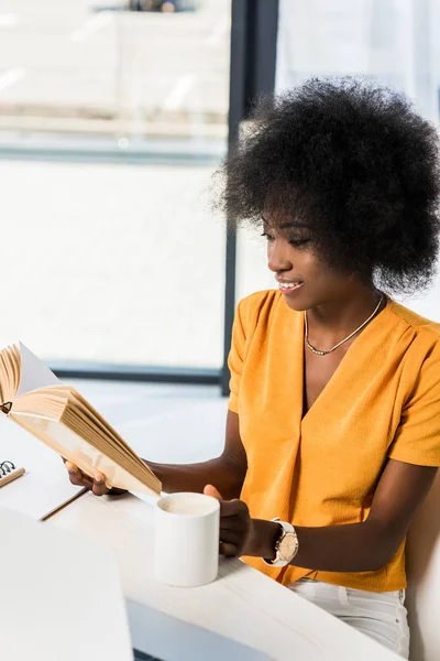 Souriant afro-américain avec tasse de livre de lecture de café à la table au bureau à domicile — Photo de stock