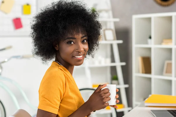 Side view of smiling african american with cup of coffee in hands at home office — Stock Photo