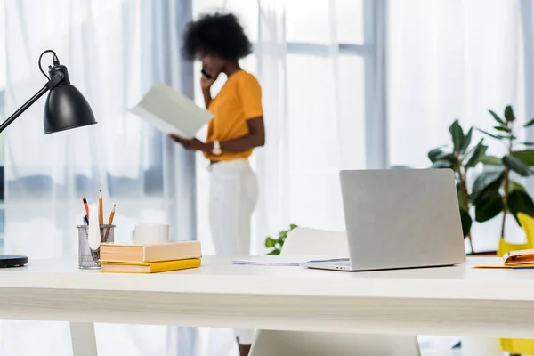 Enfoque selectivo de la computadora portátil en el lugar de trabajo y el freelancer afroamericano hablando en el teléfono inteligente en el fondo en casa - foto de stock