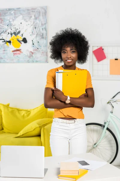 Retrato de mulher afro-americana sorridente com pastas em pé no local de trabalho em casa escritório — Fotografia de Stock