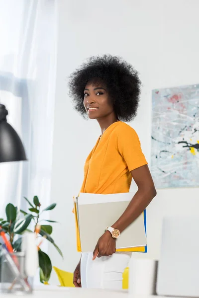 Vista lateral del sonriente freelancer afroamericano con papeles en la mano en la oficina en casa - foto de stock