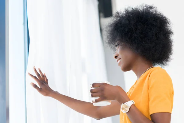 Side view of smiling african american woman with cup of coffee looking out window at home — Stock Photo
