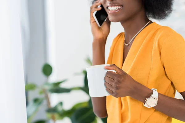Частичный вид улыбающегося африканского американца с чашкой кофе разговаривающего на смартфоне дома — стоковое фото