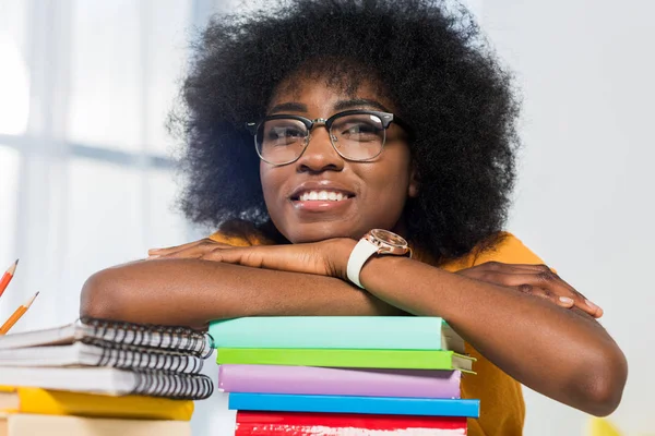 Smiling african american freelancer in eyeglasses leaning on pile of notebooks at workplace at home office — Stock Photo