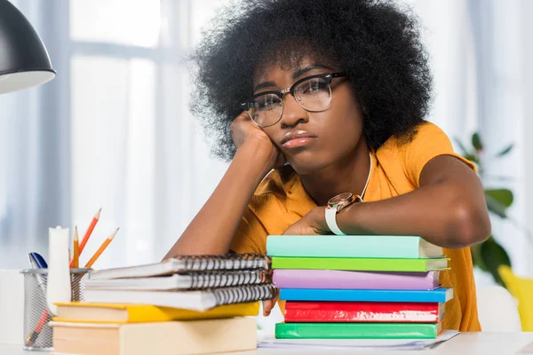 Tired african american freelancer in eyeglasses leaning on pile of notebooks at workplace at home office — Stock Photo