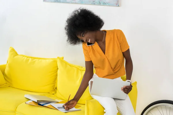 Sorrindo Africano americano feminino freelancer com laptop falando no smartphone enquanto trabalhava no sofá em casa — Fotografia de Stock