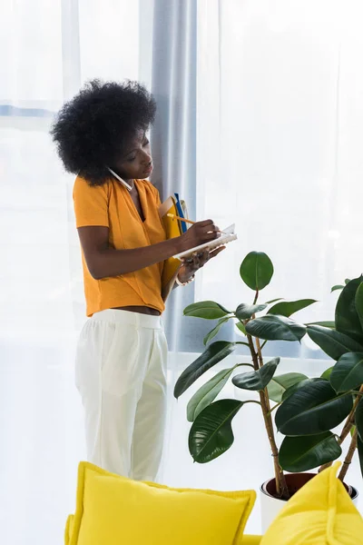 Donna afroamericana con cartelle prendere appunti nel taccuino mentre parla su smartphone a casa — Foto stock