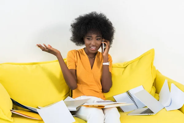 Portrait of confused african american freelancer talking on smartphone on sofa with papers at home — Stock Photo