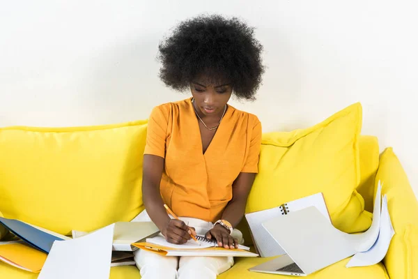 Afro-américaine pigiste prendre des notes dans un carnet tout en travaillant sur un canapé avec ordinateur portable à la maison — Photo de stock