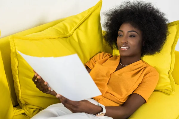 Femme afro-américaine souriante avec des papiers à distance travaillant sur un canapé à la maison — Photo de stock