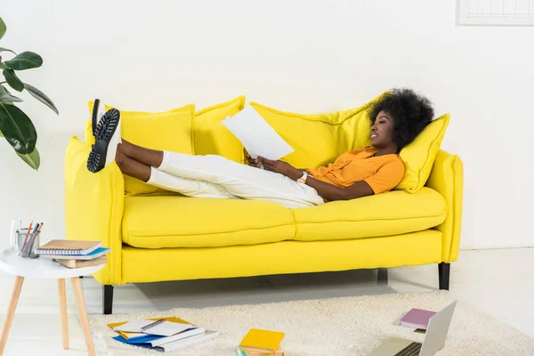 Seitenansicht einer afrikanisch-amerikanischen Frau mit Papieren, die zu Hause auf dem Sofa arbeiten — Stockfoto