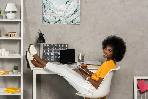 Улыбающаяся африканская американка с ноутбуком, сидящая за столом с ноутбуком в домашнем офисе — стоковое фото