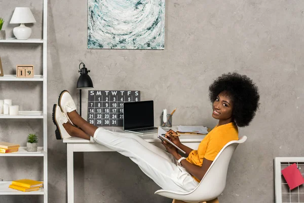 Femme afro-américaine souriante avec ordinateur portable assis à la table avec ordinateur portable au bureau à la maison — Photo de stock