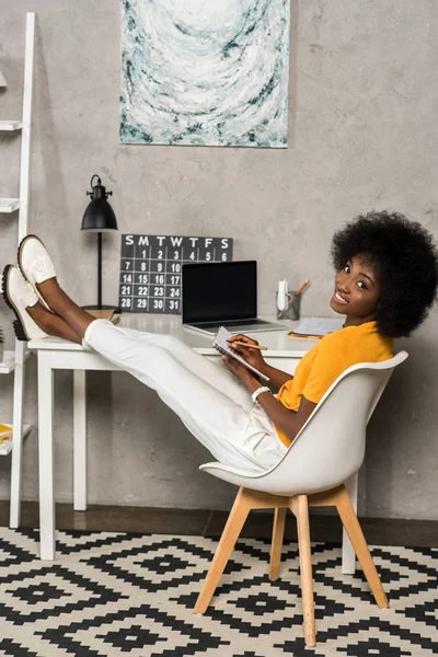 Sonriente mujer afroamericana con cuaderno sentado en la mesa con el ordenador portátil en la oficina en casa - foto de stock