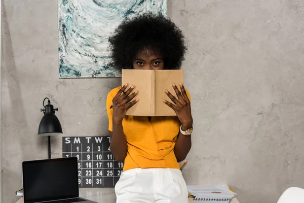 Obscured view of african american woman covering face with notebook in hands at home office — Stock Photo