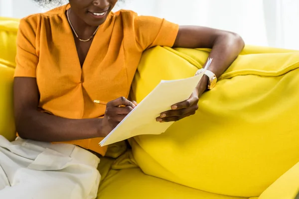 Vue partielle d'une femme afro-américaine souriante avec des papiers travaillant à distance sur un canapé à la maison — Photo de stock