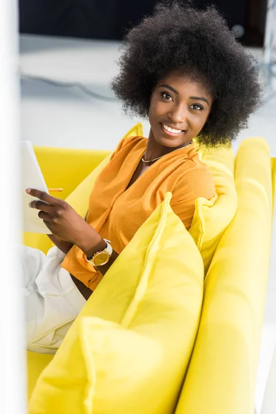 Vista lateral de la sonriente mujer afroamericana con papeles mirando a la cámara mientras trabaja a distancia en el sofá en casa - foto de stock