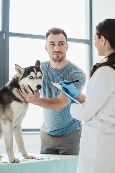 Sonriente hombre sosteniendo husky y mujer veterinaria escritura en portapapeles - foto de stock