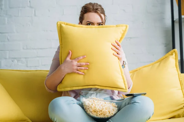 Женщина смотрит из подушки во время просмотра фильма ужасов с попкорном дома — стоковое фото
