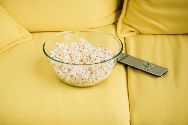 Glass bowl with popcorn and remote control on yellow sofa — Stock Photo