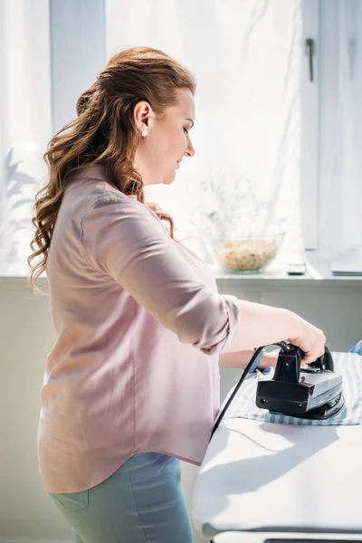 Vista laterale di bella donna camicia da stiro con ferro da stiro a casa — Foto stock