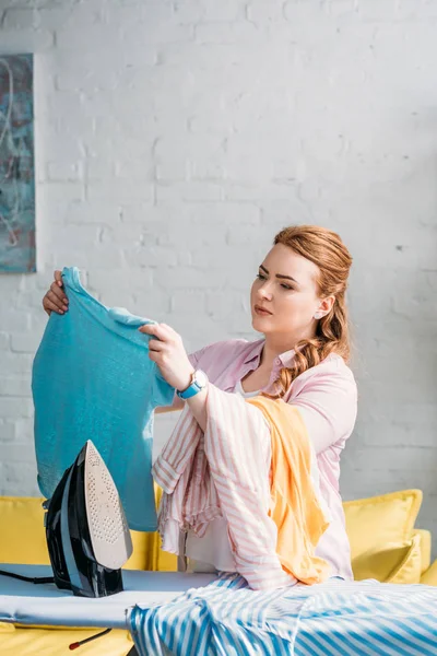 Красивая женщина смотрит на рубашку возле гладильной доски дома — стоковое фото