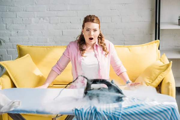 Femme choquée regardant fer brûlé avec de la fumée sur planche à repasser à la maison — Photo de stock