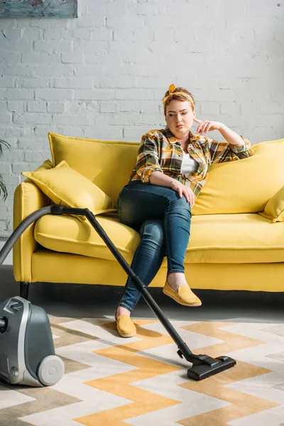 Belle femme assise sur le canapé et regardant aspirateur à la maison — Photo de stock