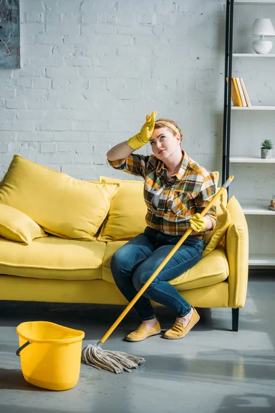 Müde Frau sitzt zu Hause mit Wischmopp auf Sofa — Stockfoto