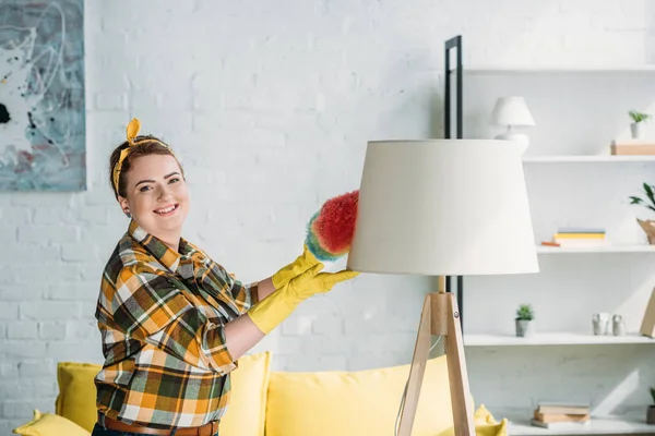 Beautiful woman dusting lamp with dust brush at home — Stock Photo