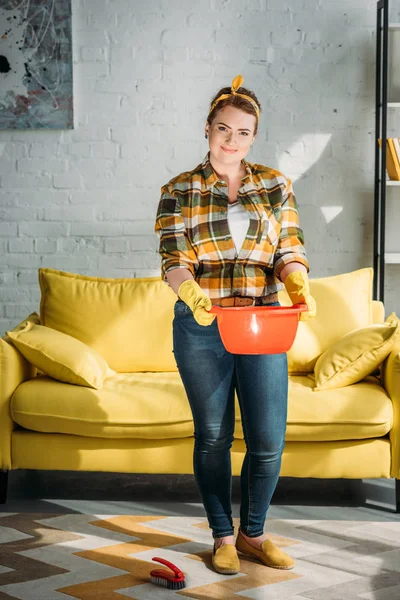 Atractiva mujer sosteniendo cubo rojo con agua para limpiar en casa - foto de stock