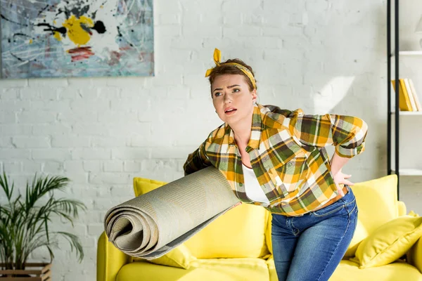 Beautiful woman carrying carpet and having pain in back at home — Stock Photo
