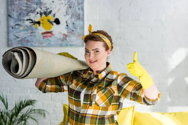 Beautiful woman holding carpet on shoulder and showing thumb up at home — Stock Photo