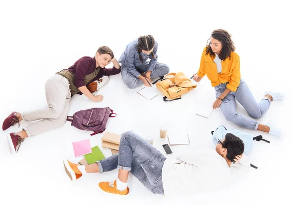 Vista de ángulo alto de estudiantes interracial con cuadernos y mochilas descansando aislados en blanco - foto de stock