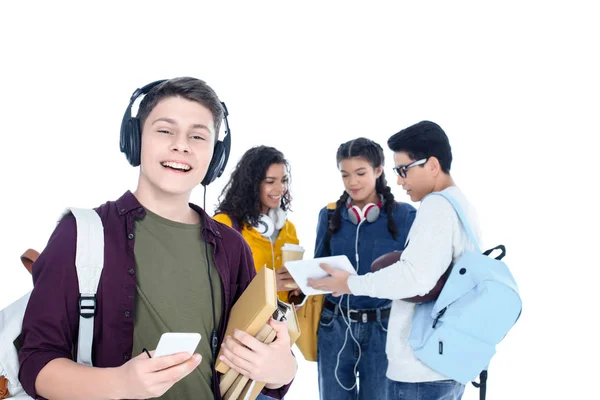 Handsome teen student in headphones and with smartphone isolated on white with his friends chatting on background — Stock Photo