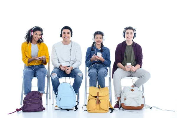 Group of teen students in headphones sitting on chairs isolated on white — Stock Photo
