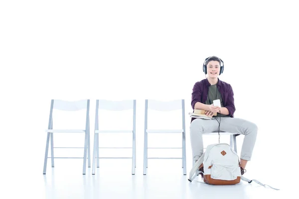 Sonriente adolescente estudiante chico escuchar música mientras sentado en silla aislado en blanco - foto de stock
