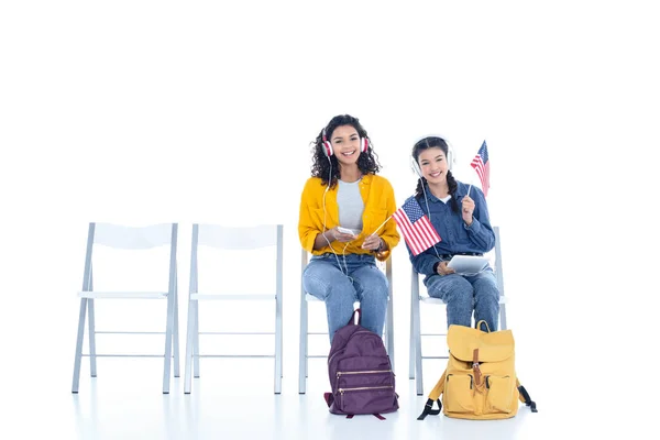 Teenager-Studentinnen mit US-Flaggen hören Musik mit Kopfhörern, während sie isoliert auf weißen Stühlen sitzen — Stockfoto