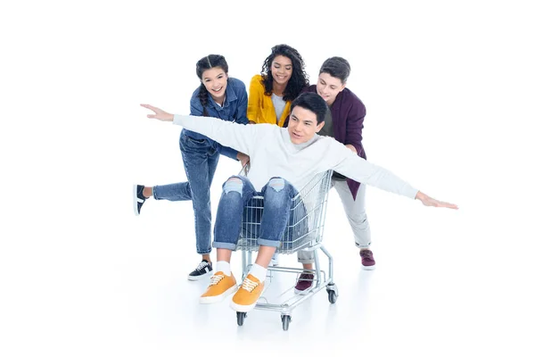 Happy teen students riding their friend on shopping cart isolated on white — Stock Photo